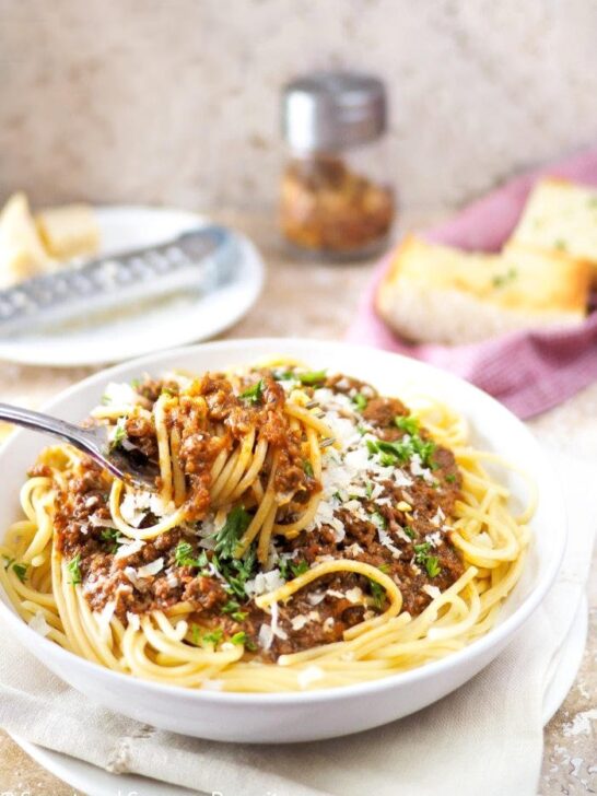 Close-up of spaghetti and meat sauce twirled around a fork. Garlic bread in background.