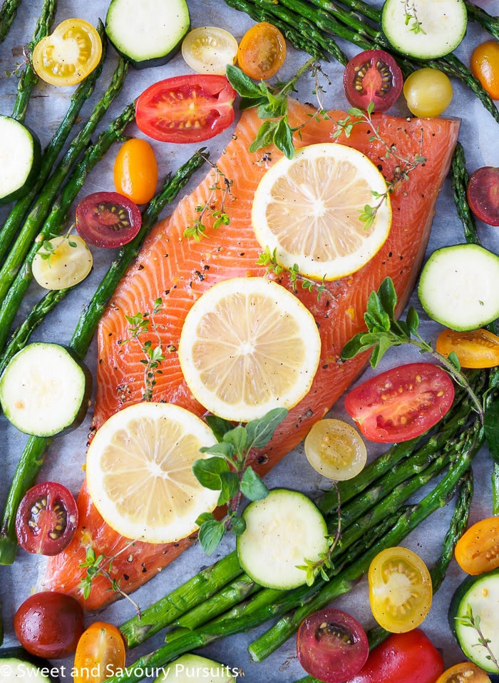 Unbaked rainbow trout fillet on baking sheet with asparagus, cherry tomatoes and zucchini with fresh herbs.