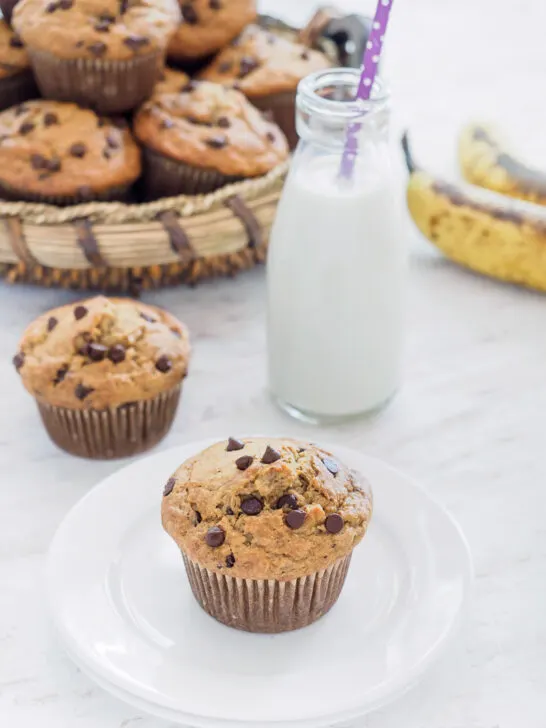 A basket of Banana Oatmeal Chocolate Chip Muffins and one muffin on small dish served with milk.