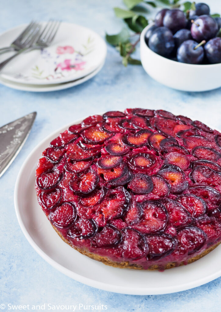 Plum Upside Down Cake on white dish with fresh plums in background.