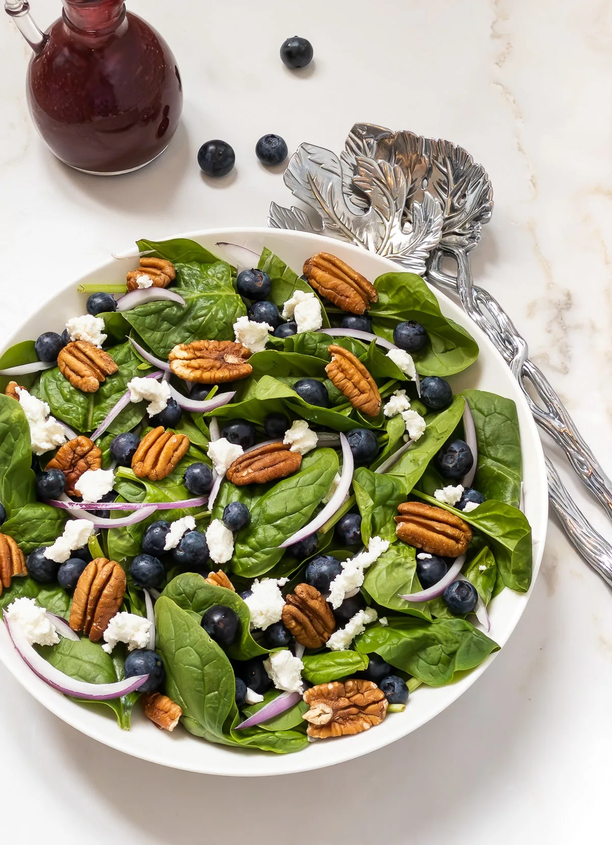 Large bowl of spinach and blueberry salad topped with toasted pecans, goat cheese and slivers of red onion.