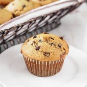Banana chocolate chip muffin on small dish with basket of muffins in background.