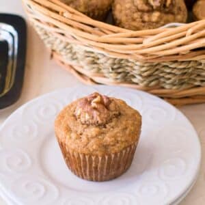 Banana muffin on small white dish with basket of muffins in the background.